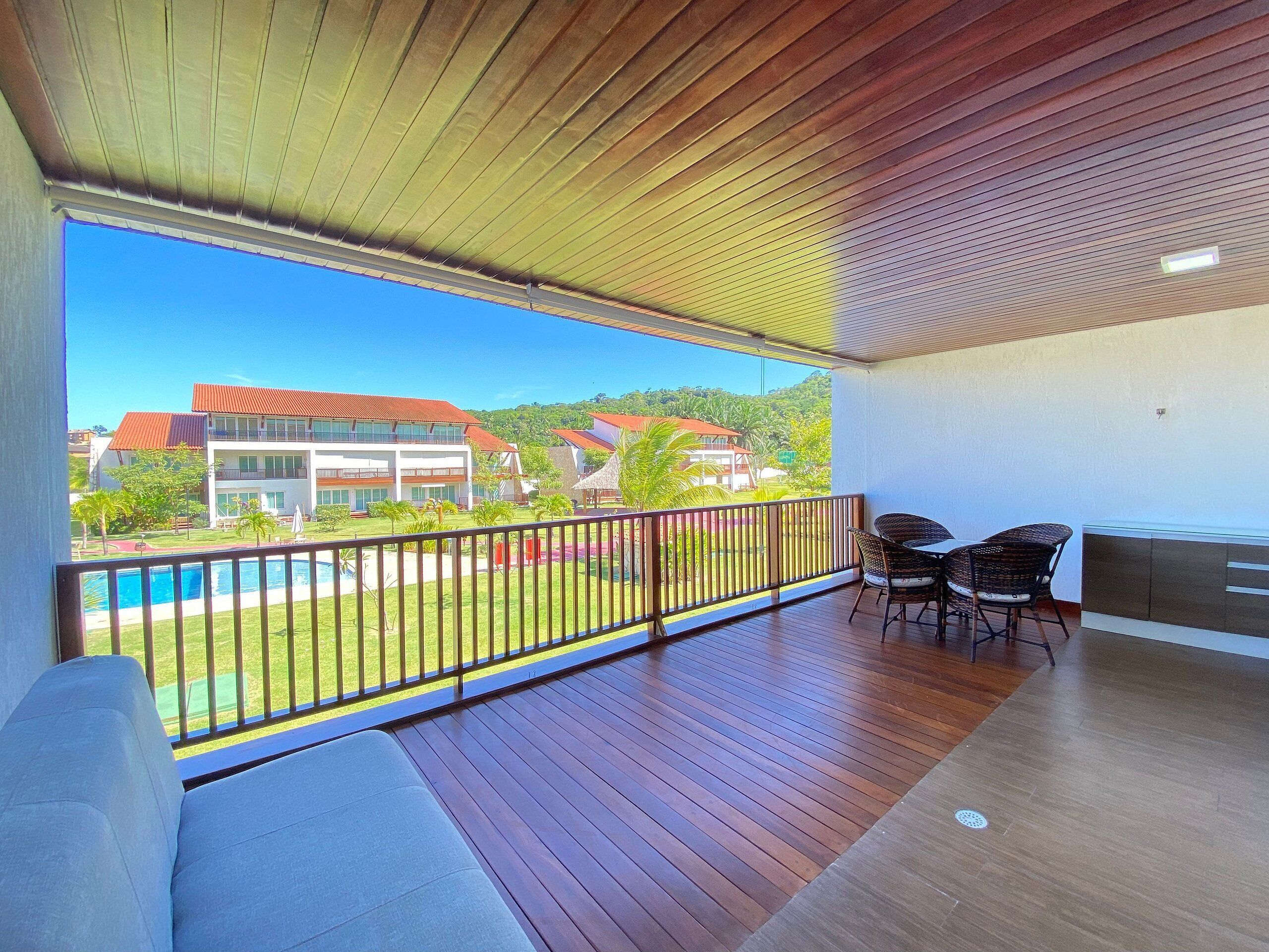 A balcony with a view of a swimming pool and buildings, wooden flooring, and a seating area with chairs.