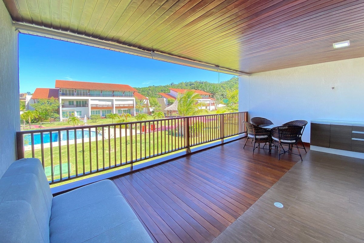 A balcony with a view of a swimming pool and buildings, wooden flooring, and a seating area with chairs.