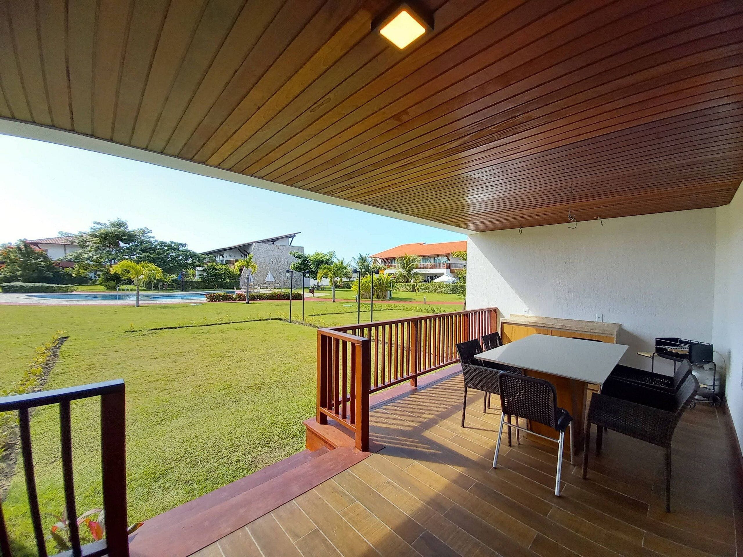 A covered outdoor area with a table and chairs, a wooden railing, and a view of a lawn and buildings in the background.