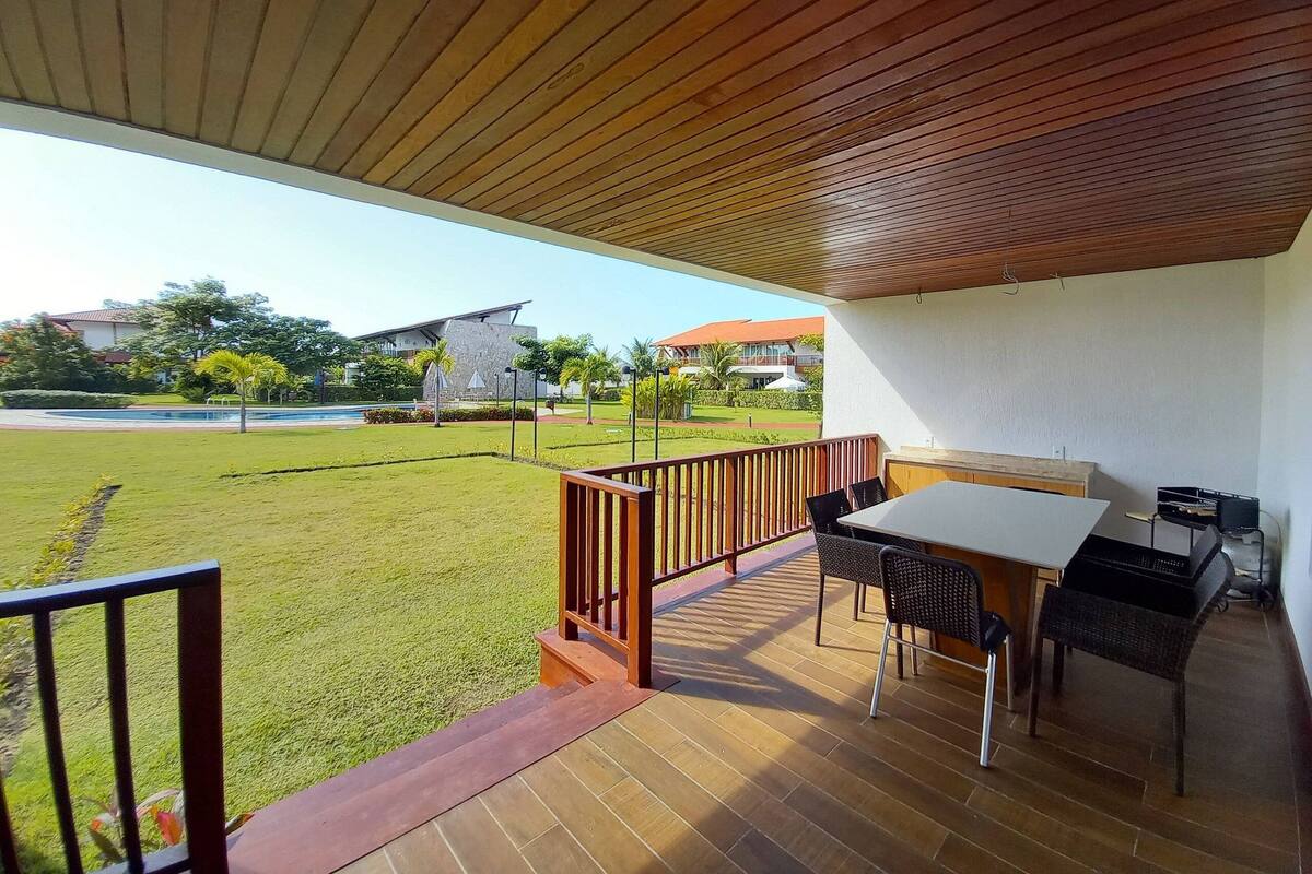 A covered outdoor area with a table and chairs, a wooden railing, and a view of a lawn and buildings in the background.