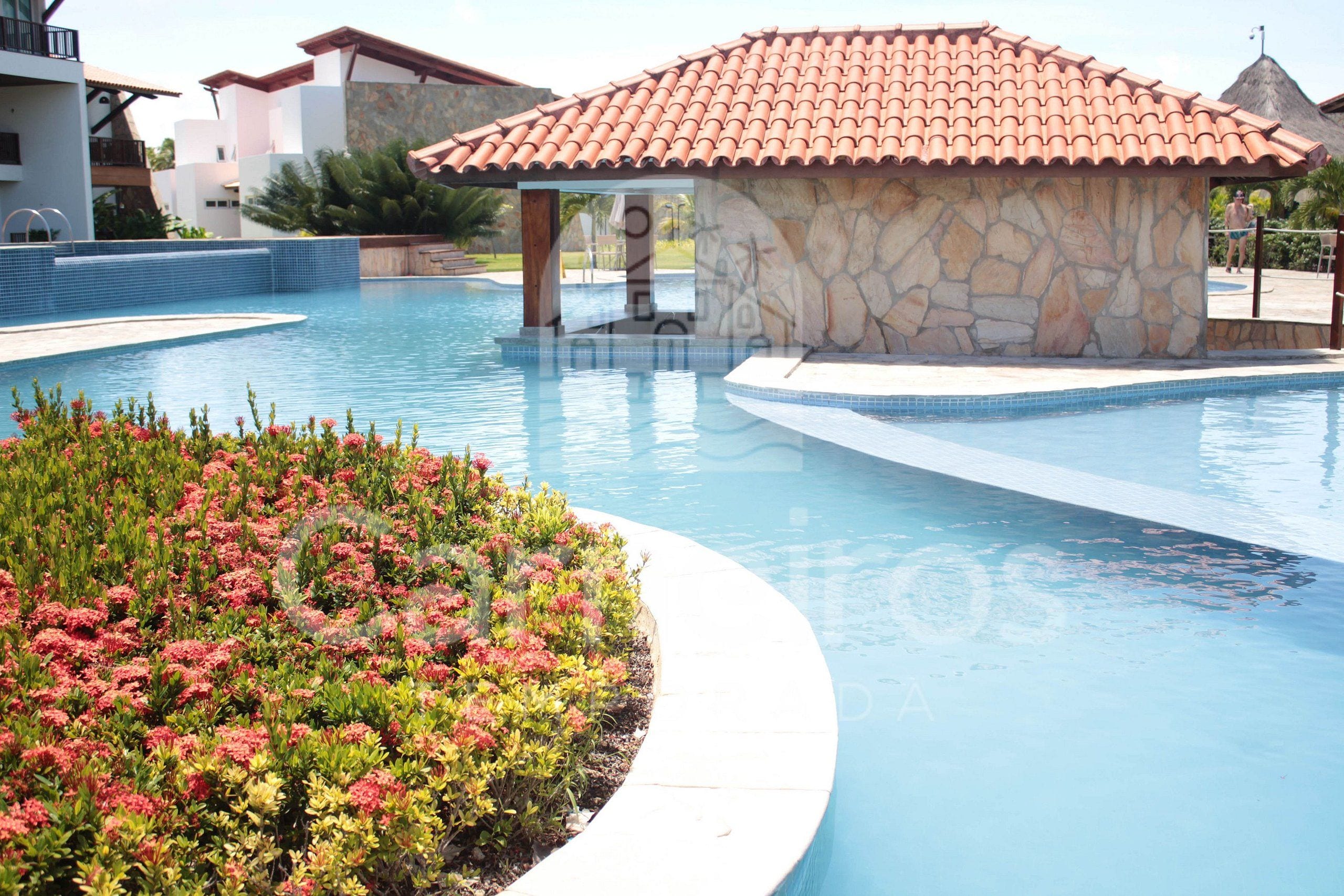 Une piscine dotée d’une structure en pierre, entourée d’un parterre de fleurs et bordée d’un bâtiment au toit recouvert de tuiles.