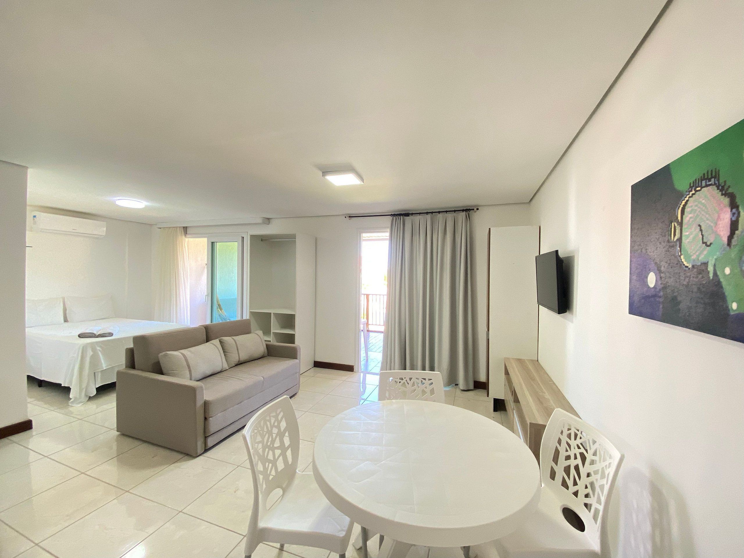 A hotel room with a bed, a sofa, a dining table with chairs, a TV, and a painting on the wall.