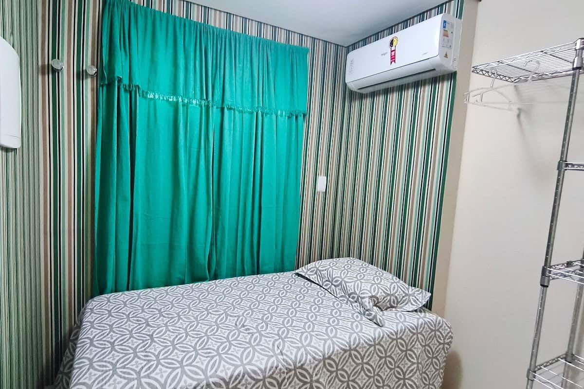 A room with a bed, striped curtains, an air conditioning unit, and a metal shelf.