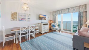 A room with a wooden floor, a desk with chairs, a sofa, a TV, and a view of the sea through large windows. - North Myrtle Beach Oceanfront Condo w Lazy River (North Myrtle Beach)
