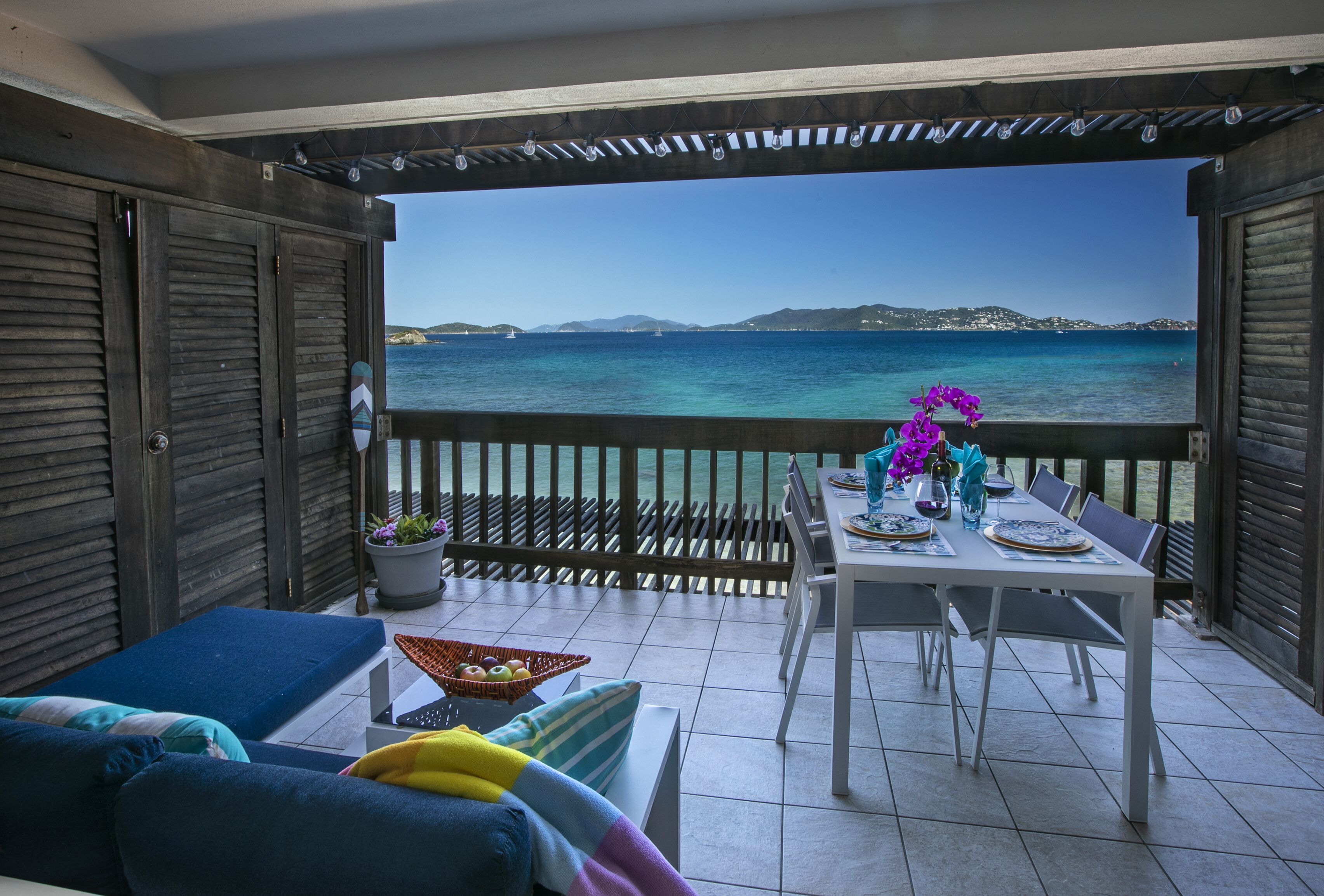 Uma varanda com vista para o mar, uma mesa de jantar posta com pratos e copos, e um sofá com almofadas.
