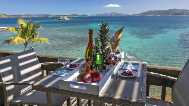 A table set with fruits, bottles, and glasses on a balcony with a view of the ocean.