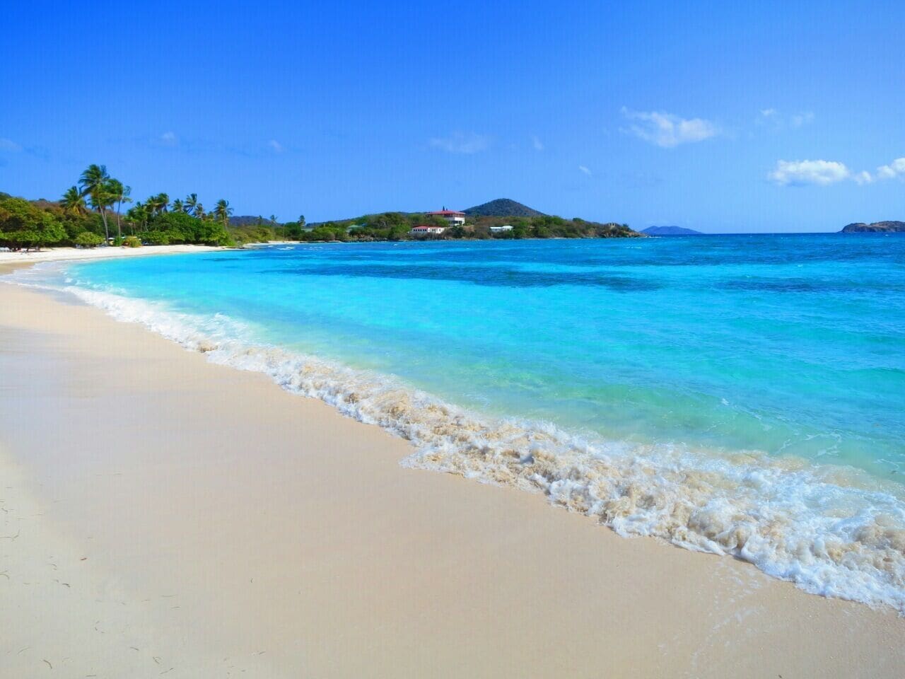 A beach with clear turquoise water, a sandy shore, and a small island with buildings in the background.