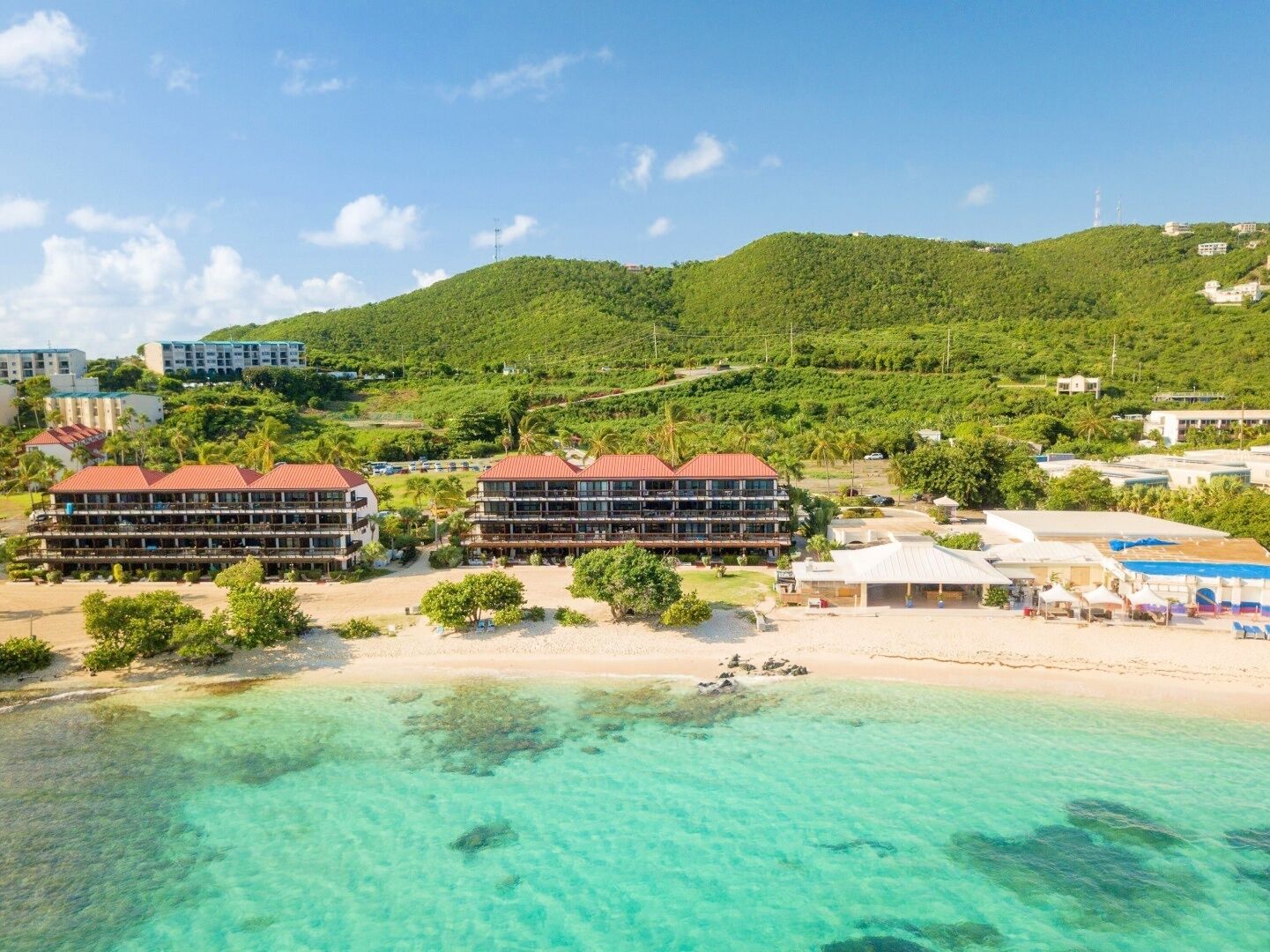 Un hotel a la costa amb diversos edificis, una platja i aigües turqueses cristal·lines.
