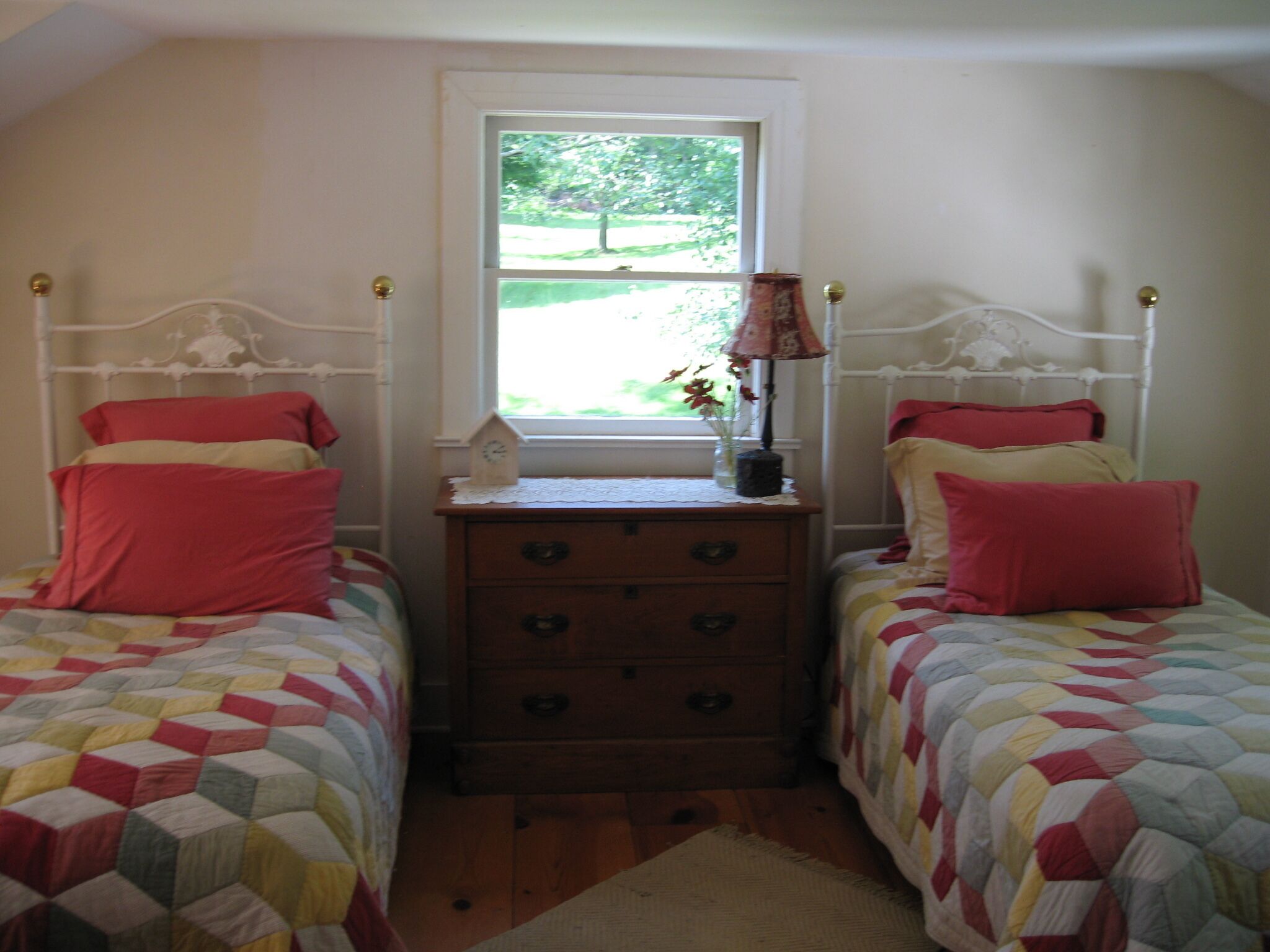 Une chambre avec deux lits simples, une commode en bois, une fenêtre donnant sur un paysage verdoyant et une lampe sur la commode.