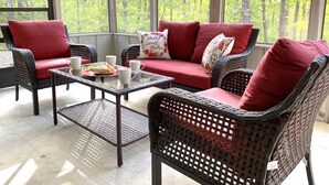 Une terrasse ensoleillée, meublée de meubles en osier, avec une table en verre et un canapé rouge agrémenté de coussins à motifs floraux.