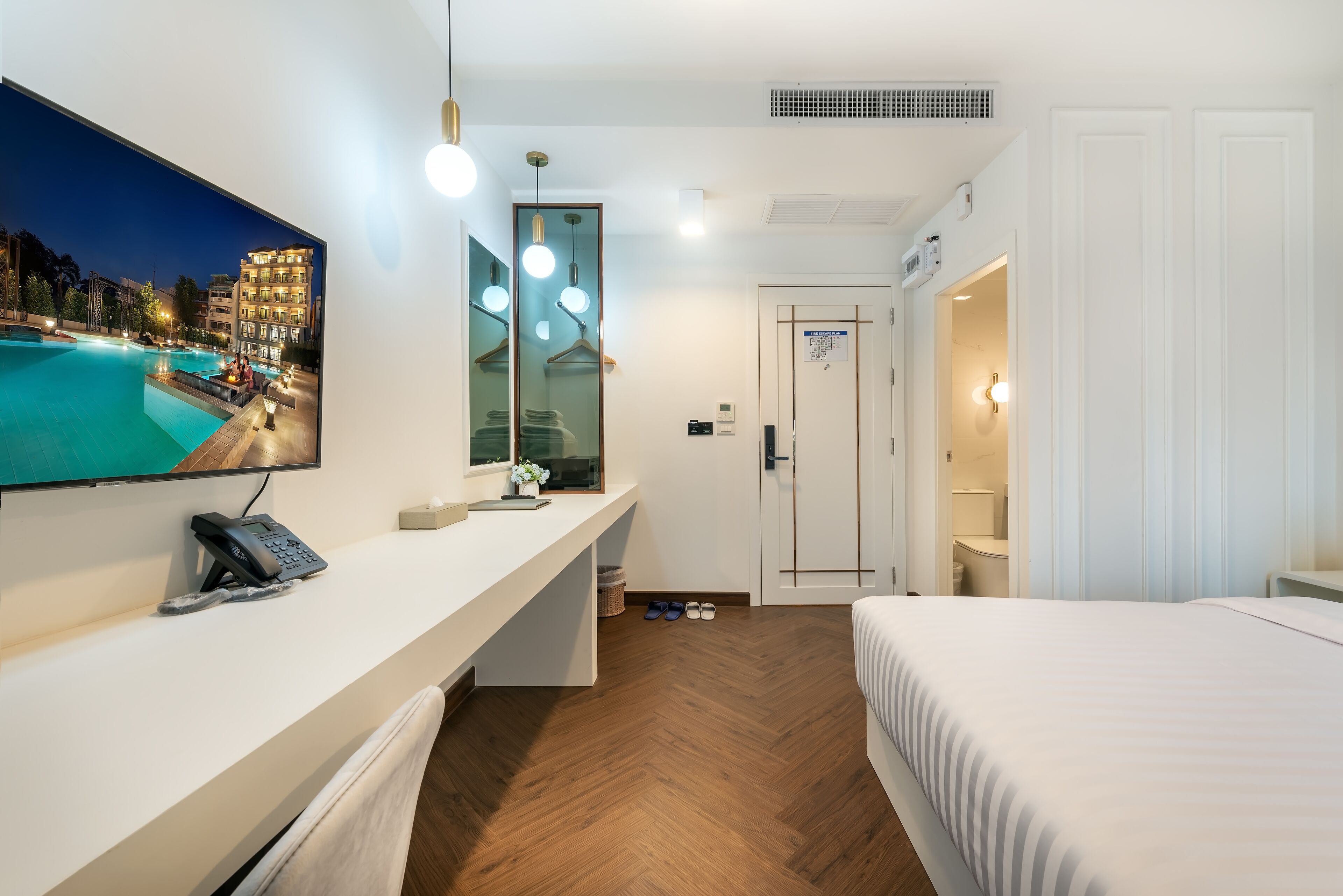 Habitación de hotel con televisor grande mostrando vista a la piscina, escritorio con teléfono, espejo y puerta que da a un baño.