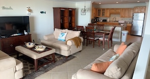 Ein Wohnzimmer mit einer Couch, einem Couchtisch und einem Fernseher. Ein Essbereich mit Tisch und Stühlen ist ebenfalls vorhanden. Im Hintergrund ist eine Küche mit Holzausstattung und einem Kühlschrank zu sehen.