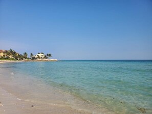 A beach with clear turquoise water, a sandy shore, and a building with a white facade and palm trees in the background. - Gorgeous 3BR Oceanfront Villa - Amazing Views of Turquoise Water (Freeport)