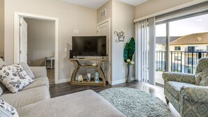 Una sala de estar con un televisor sobre un mueble de madera, un sofá, un sillón estampado y un amplio ventanal con vistas a edificios.
