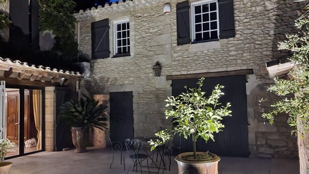 A stone building with a courtyard, featuring a potted tree, outdoor seating, and a large pot with plants. - LE MAS DES COSME EN PROVENCE (Aramon)