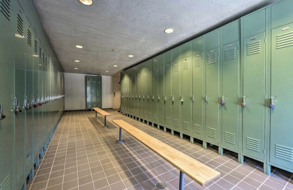 A locker room with green lockers, a wooden bench, and a tiled floor.