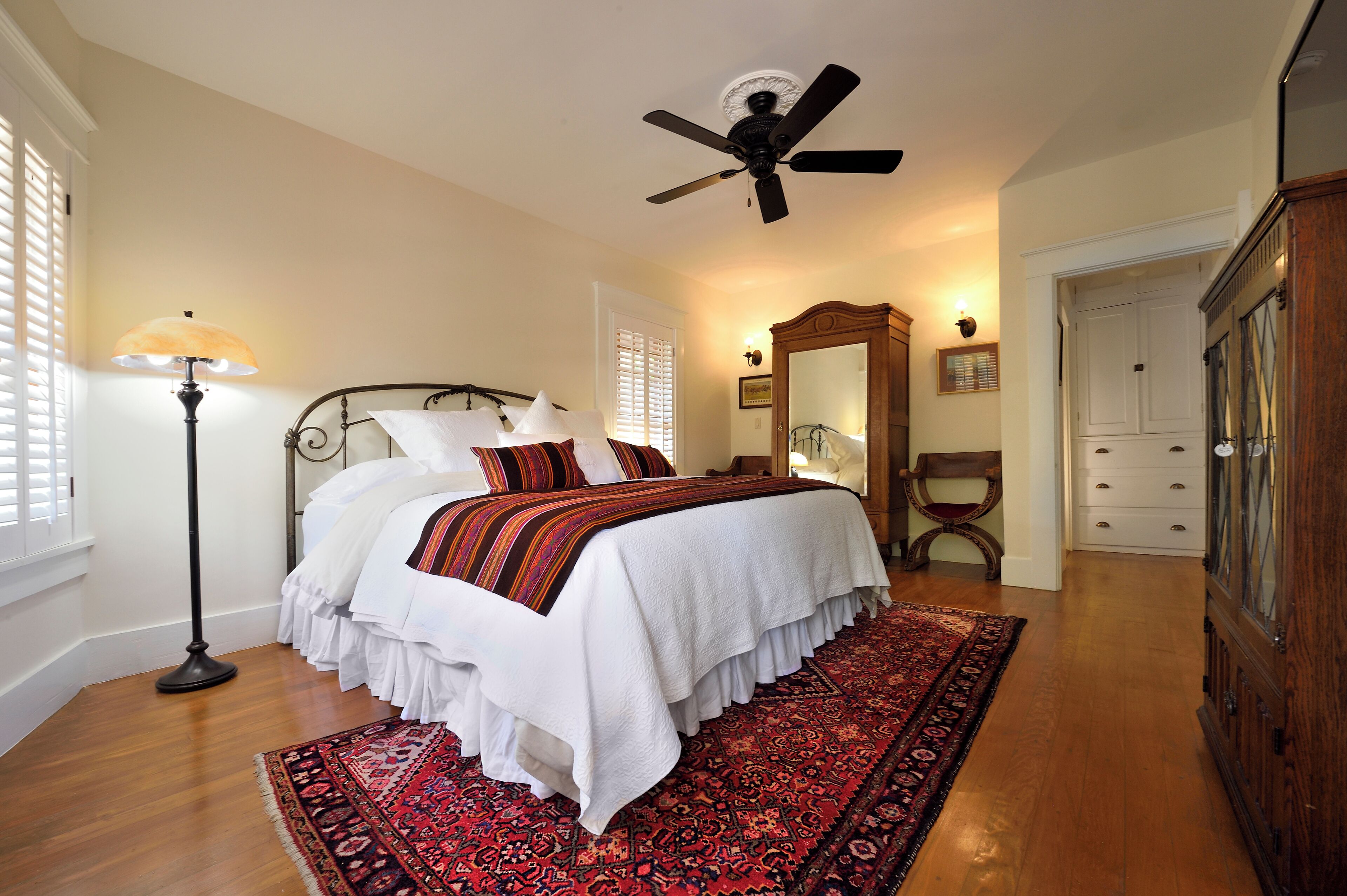 A bedroom with a bed, a standing lamp, a ceiling fan, a wooden dresser, and a patterned rug.