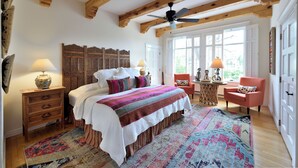 Ein Schlafzimmer mit einem großen Bett, Holzbalken, einem Deckenventilator, einem Fenster mit Rollläden, einem kleinen Tisch mit Lampe und einem Sessel.