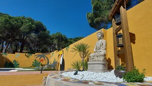 Uma estátua de uma figura sentada em um jardim, com uma parede amarela, piso de azulejos e uma pérgola de madeira com lanternas.