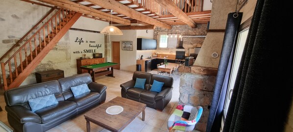 Amplia sala de estar con una escalera de madera, sofás de cuero, mesa de billar y una cocina con paredes de piedra.