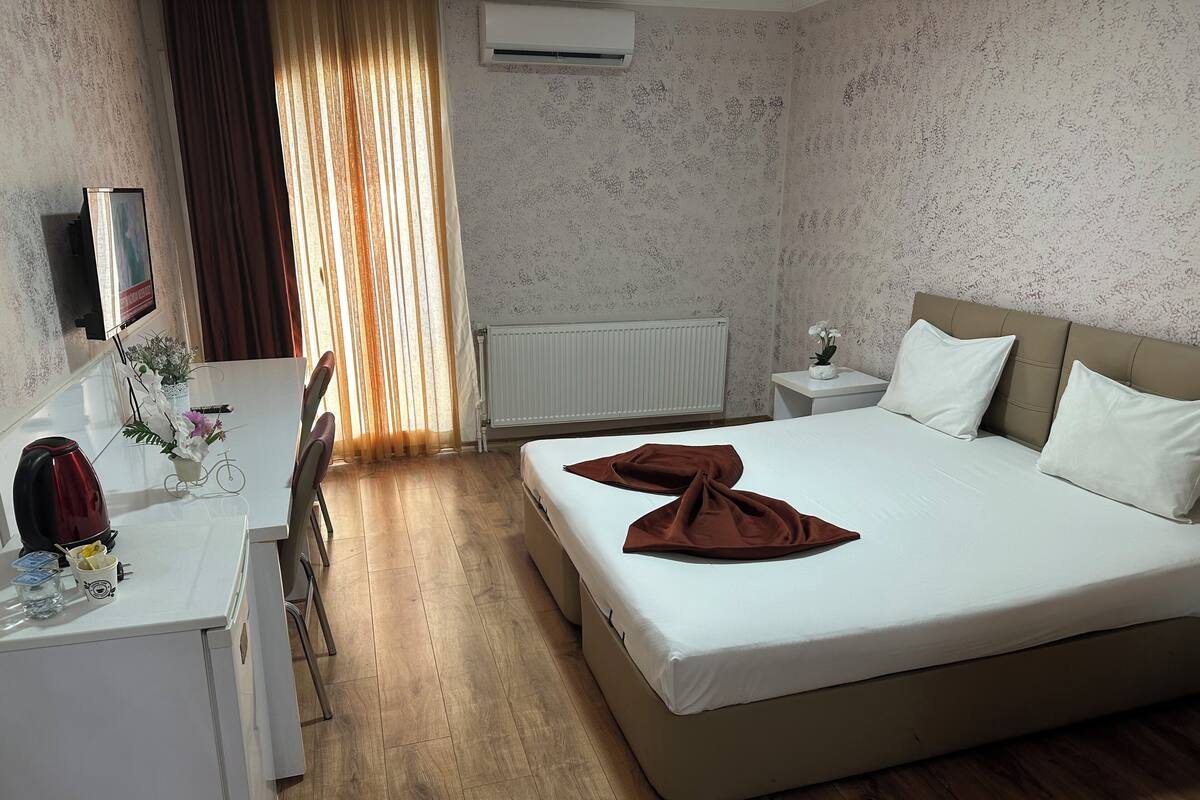 A hotel room with a bed, a desk, a chair, a radiator, and a window with curtains.