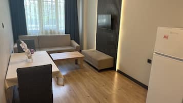 A modern living room with a sofa, a coffee table, a dining table with chairs, a flat-screen TV mounted on the wall, and a refrigerator.