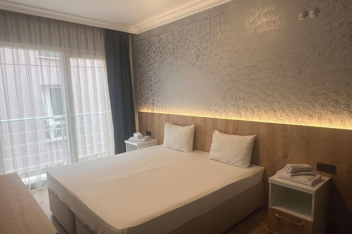Ein großes Bett mit Holz-Kopfteil, ein Nachttisch mit Lampe und Blick durch große Fenster nach draußen.
