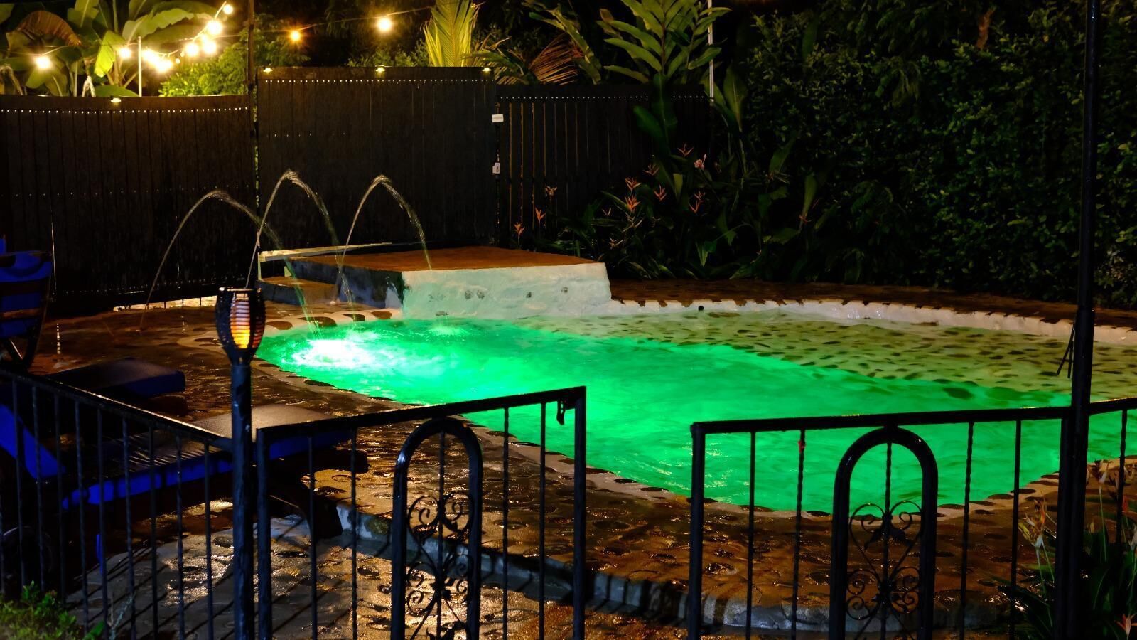 A swimming pool with a green light, surrounded by a fence and plants.