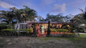 Une maison à deux étages, dotée d’un toit en tuiles, d’un porche couvert et d’un jardin bien entretenu avec des palmiers et des arbustes.