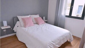 A neatly made bed with white and pink bedding, a bedside table with a lamp, and a window with a view of buildings outside. - Cosy, fully-equipped, refurbished F2 (Vert-le-Grand)