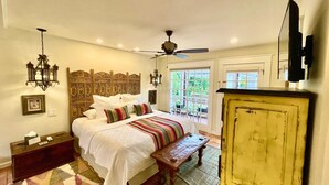 Une chambre avec un grand lit, une tête de lit en bois, un ventilateur de plafond, un banc en bois, un téléviseur et une vue sur l’extérieur par une porte.