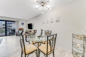 A dining area with a glass table and metal chairs, a ceiling light fixture, and a wall-mounted TV. - Cozy Beachfront Retreat; Updated modern décor (Ormond Beach)