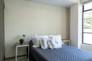 A bedroom with a bed, white pillows, a small table with a lamp, and a window.