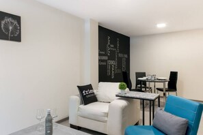 A modern, minimalist room with a white sofa, a blue armchair, and a dining table with chairs. There is a black wall with city names and a small potted plant on the table.