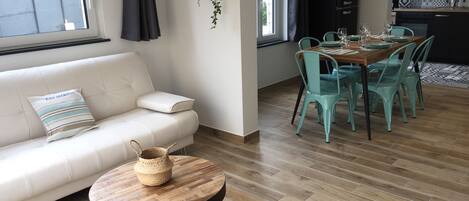 A modern living room with a white sofa, a round wooden coffee table, and a dining area with teal chairs and a wooden table.