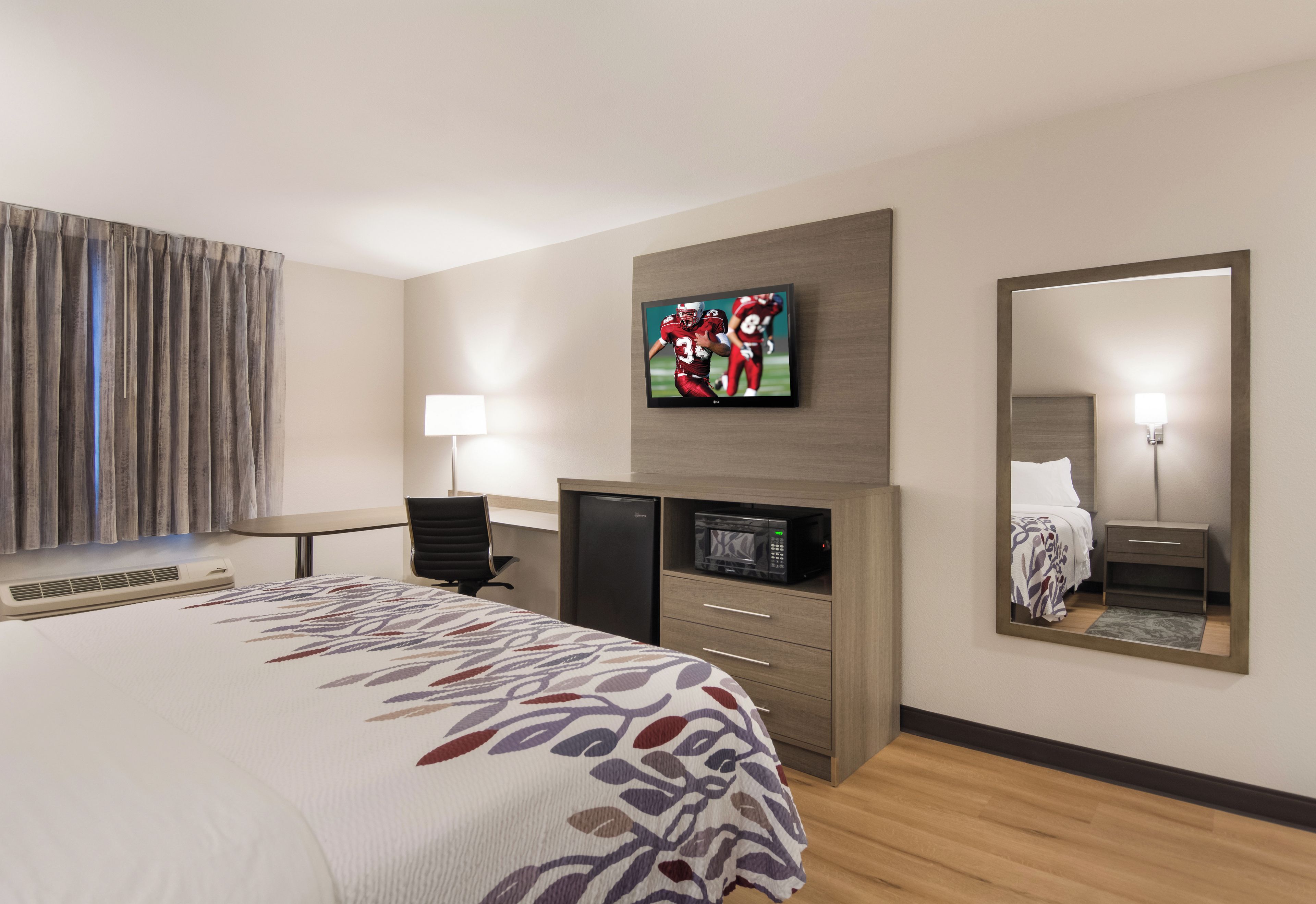 Una habitación de hotel con una cama, un televisor montado en la pared, un escritorio con una silla, un refrigerador, un microondas y un espejo.