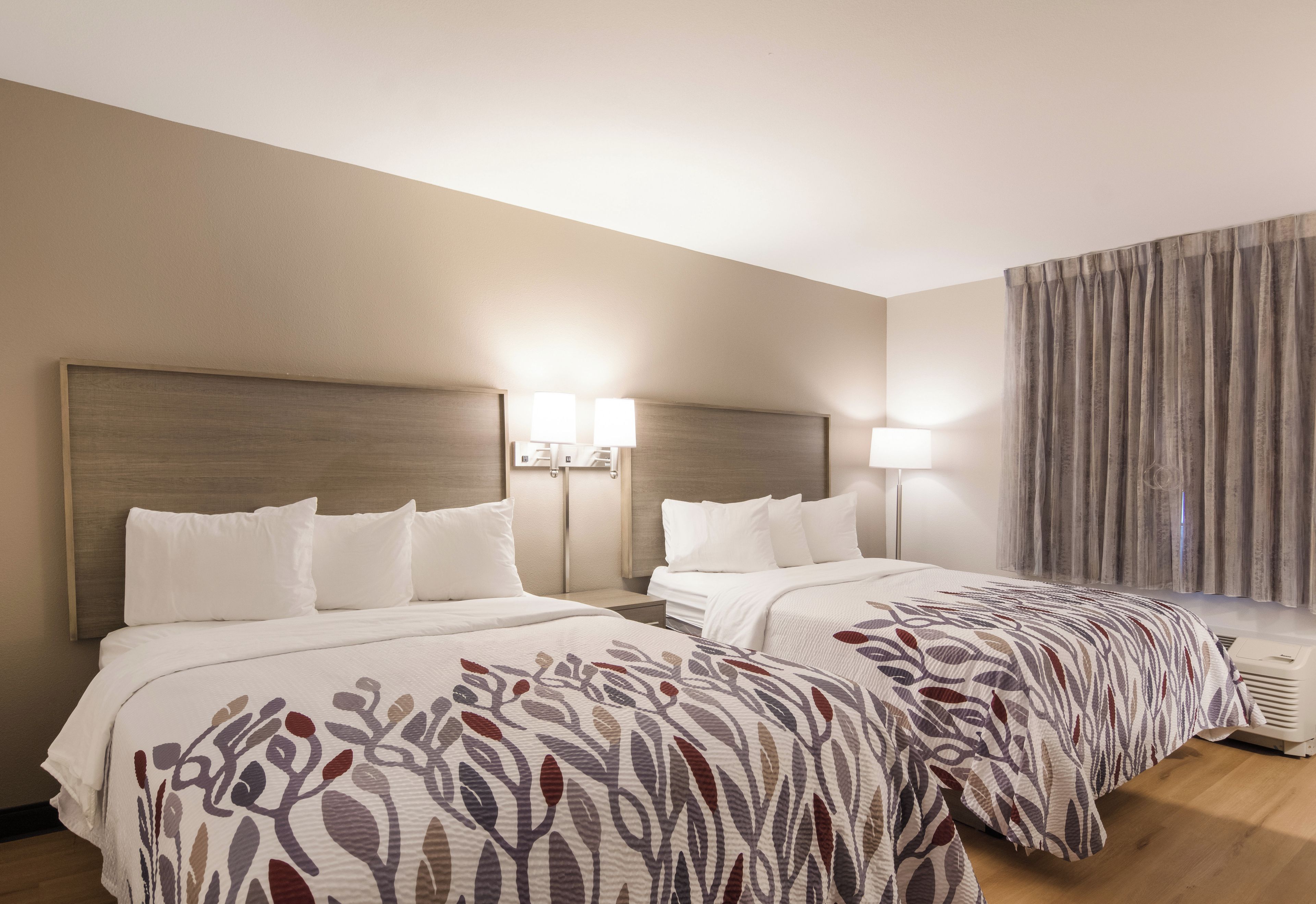 A hotel room with two beds, a headboard, bedside lamps, a window with curtains, and an air conditioning unit.