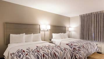A hotel room with two beds, a headboard, bedside lamps, a window with curtains, and an air conditioning unit.