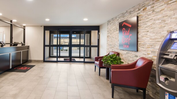 Un espace d’accueil avec un comptoir, deux chaises rouges, une plante en pot, un distributeur automatique de billets et une enseigne indiquant « Red Roof Inn ».