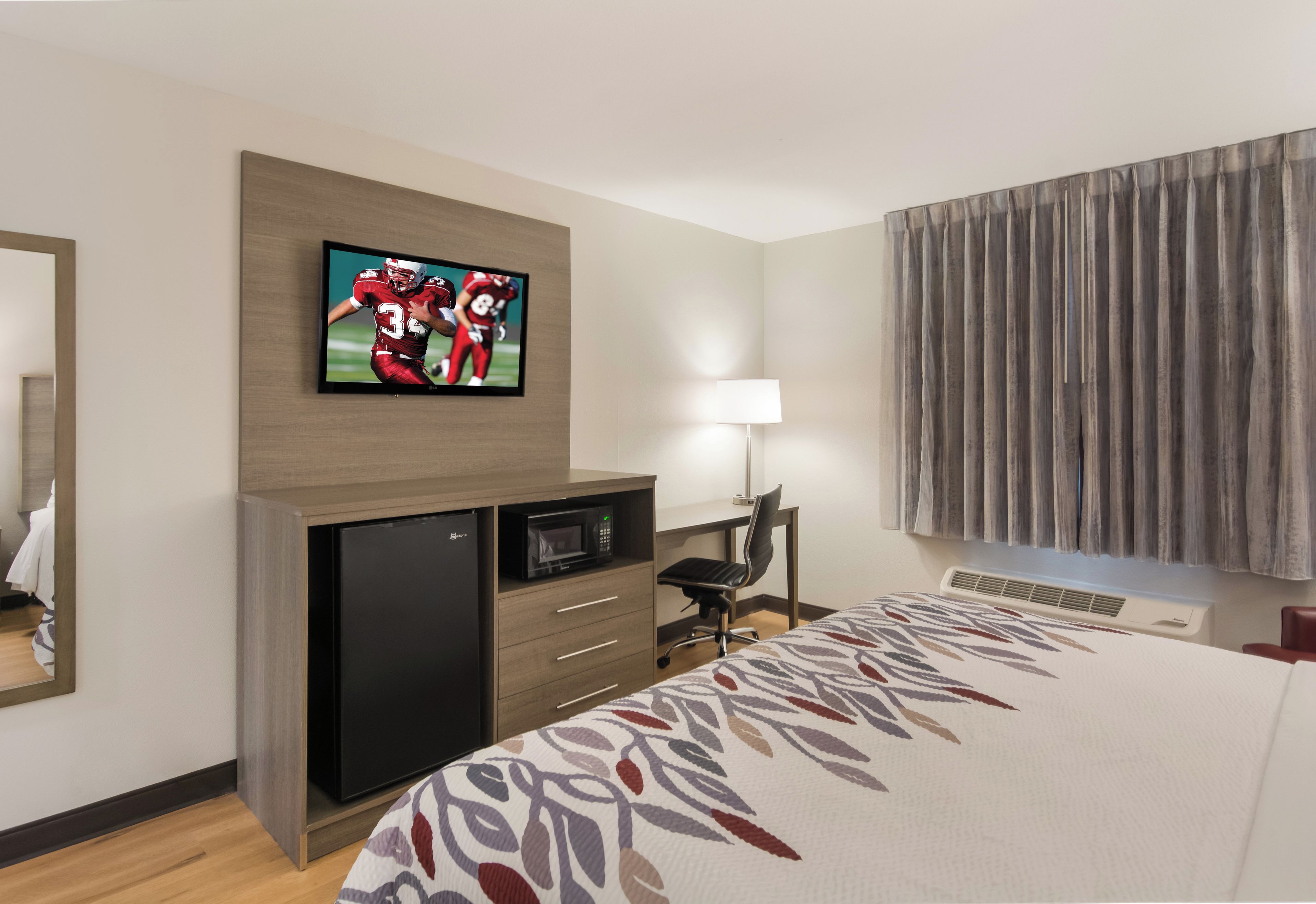 A hotel room with a bed, a desk with a computer, a television mounted on the wall, and a window with curtains.