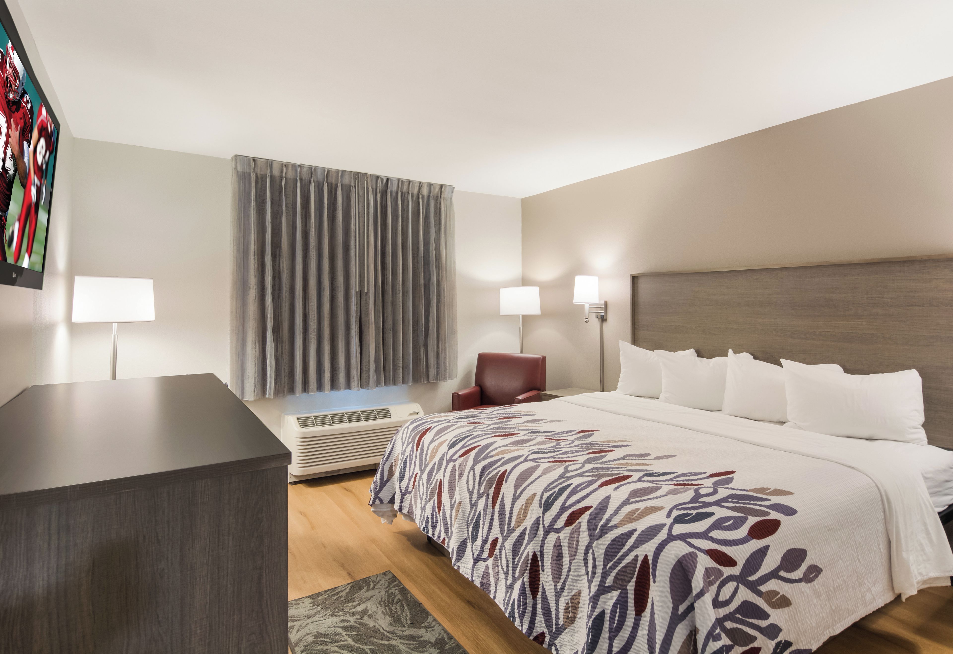 A hotel room with a bed, a desk, a chair, a TV, and a window with curtains.