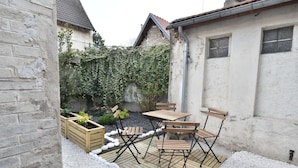 Un patio con mesa y sillas de madera, rodeado de guijarros blancos y un jardín con plantas y una enrejada.