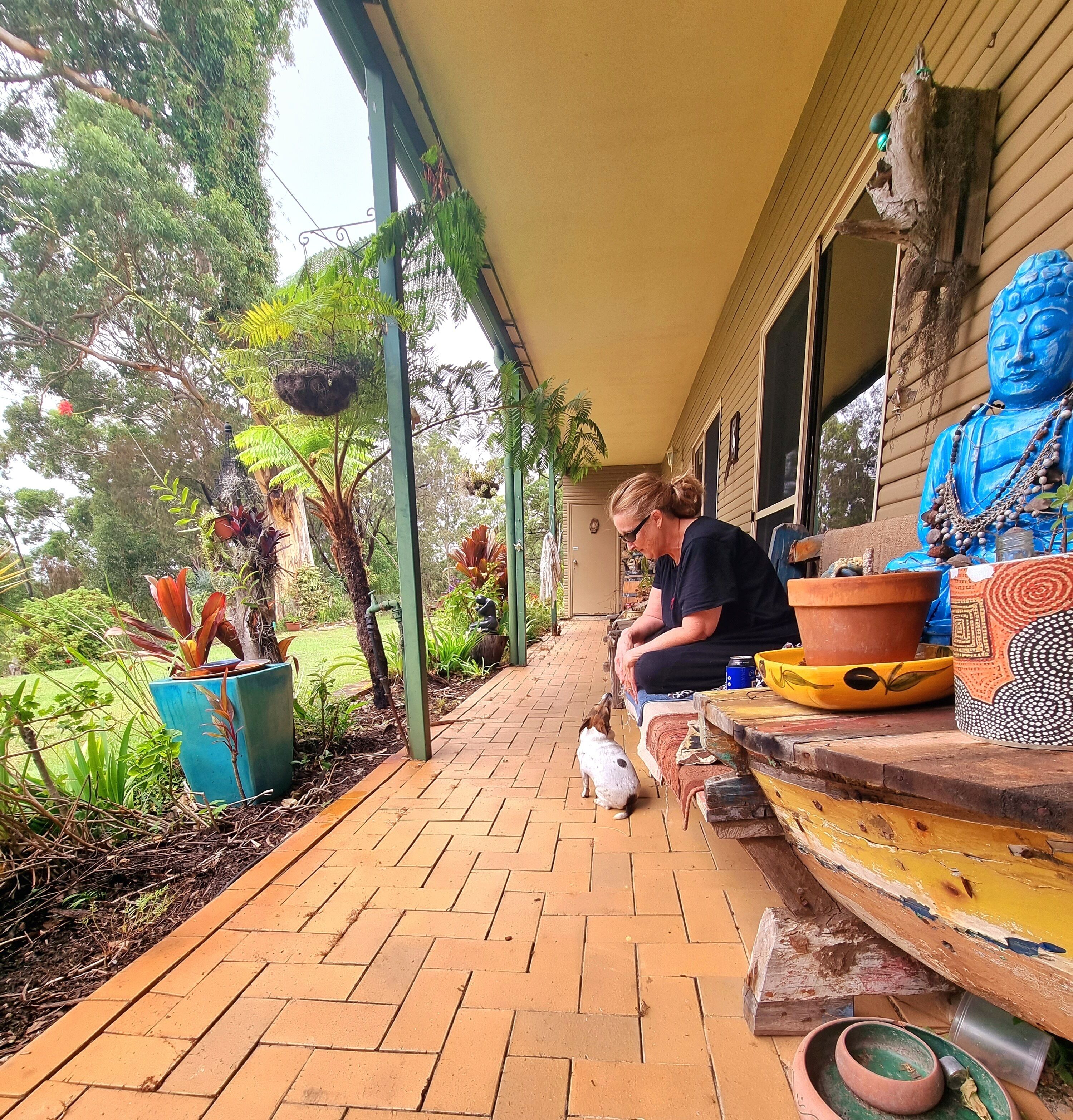 A covered porch with a person sitting on a bench, surrounded by potted plants and a Buddha statue.