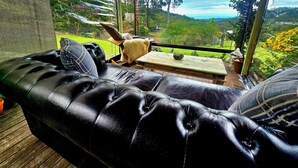 Ein überdachter Außenbereich mit einer schwarzen Ledersofa, einem Holztisch und Blick auf einen üppigen Garten und ferne Hügel.