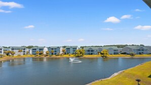 Blick auf einen See mit modernen Gebäuden, einem Brunnen und einer gepflegten Grünfläche.