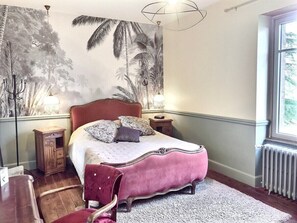 Ein Schlafzimmer mit einem großen Bett, einem Nachttisch, einer Kommode, einem Stuhl und einem Fenster mit Blick auf Bäume.