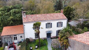 A two-story house with a red-tiled roof, a small attached garage, and a garden with palm trees and shrubs.