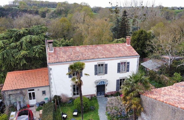 Une maison à deux étages, avec un toit recouvert de tuiles rouges, un petit garage attenant et un jardin agrémenté de palmiers et d’arbustes.