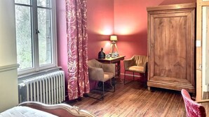 A room with a large window, a radiator, a table with a lamp, two chairs, and a wardrobe.