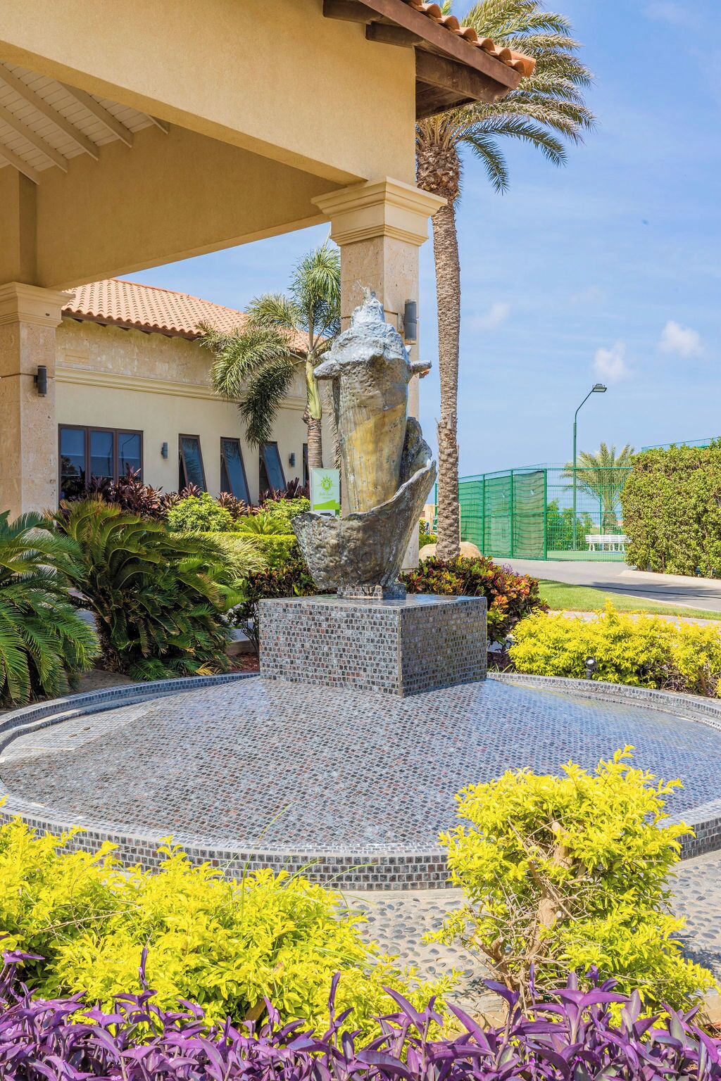 Une cour agrémentée d’une fontaine décorative, entourée de différentes plantes et bordée d’un bâtiment dont le toit est recouvert de tuiles.
